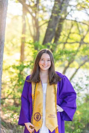 Graduate in purple academic regalia with gold honors stole poses outdoors in natural sunlit setting.