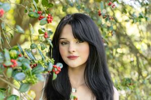 Portrait of a dark-haired woman near red berries and green foliage in natural lighting.
