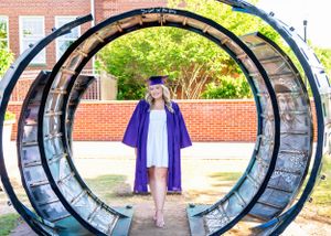 Graduate in purple gown posing under circular metal archway on campus.