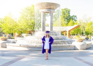 Graduate in purple cap and gown standing in front of outdoor fountain.