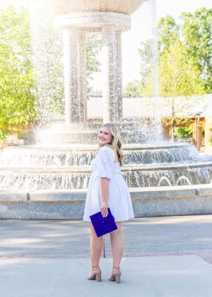 Graduate in white dress with purple accents poses by tiered fountain.