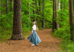 A figure in a flowing blue dress walks along a winding forest path surrounded by tall pine trees and lush greenery.