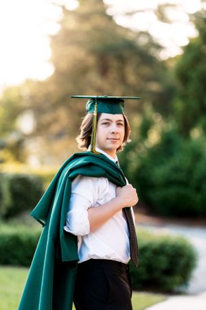 Graduate in emerald green cap and gown poses outdoors in golden sunlight.