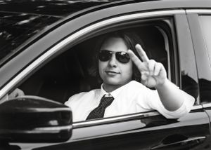 Person in sunglasses and tie making peace sign while sitting in car.