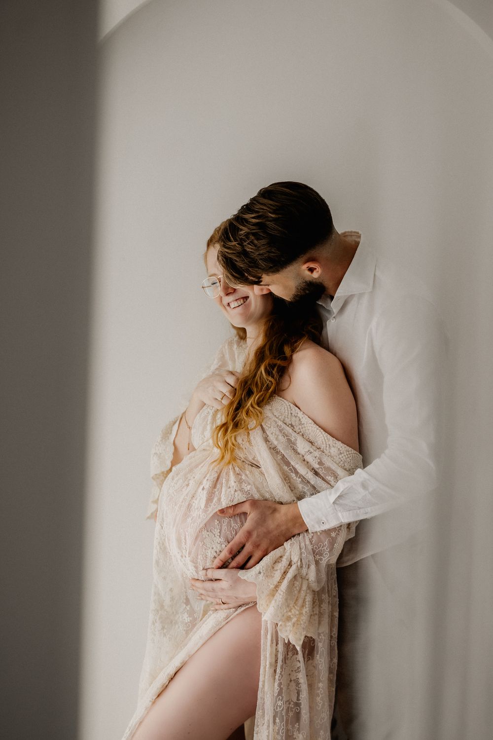 Intimate maternity photoshoot in soft light featuring flowing lace robe and gentle embrace against neutral backdrop.