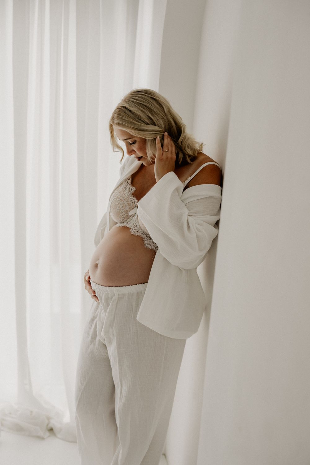 Pregnant woman in white lace bralette and linen pants cradling baby bump against a soft white wall.