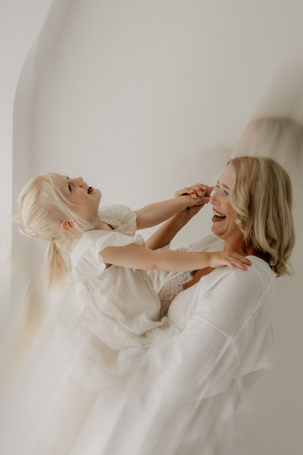 A joyful blonde mother and young daughter in white dresses laughing and playing together against a white wall.