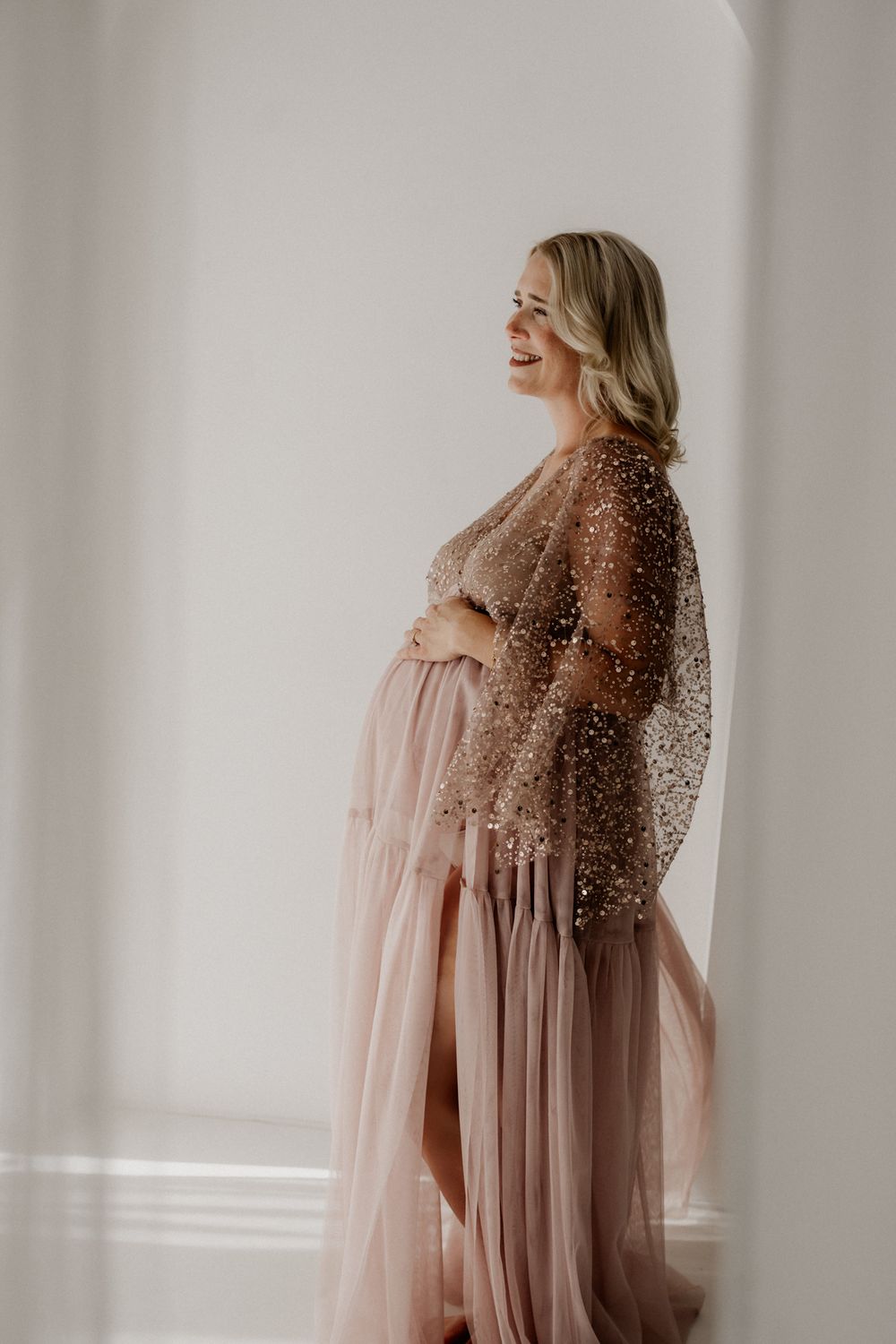 Pregnant woman in a blush pink tulle gown with sheer embellished overlay, cradling her baby bump in a bright studio.