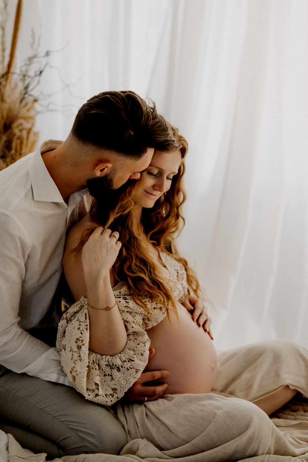 Romantic maternity photography session with couple in soft natural light against white curtain backdrop.