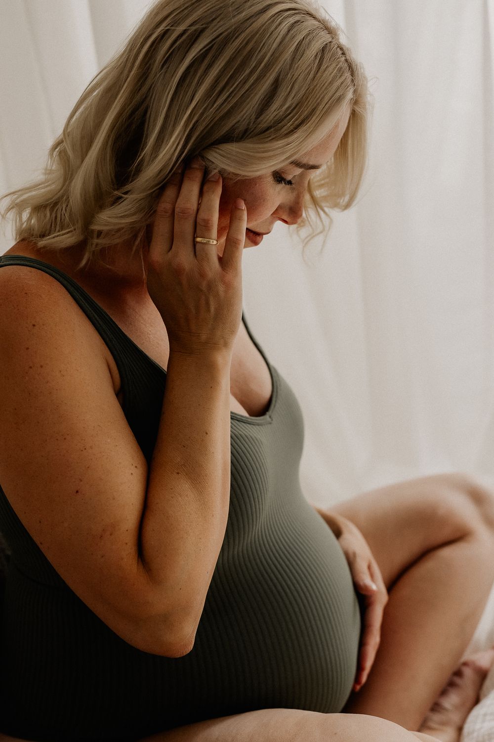 Pregnant woman in olive bodysuit gently cradling her baby bump while looking down thoughtfully.