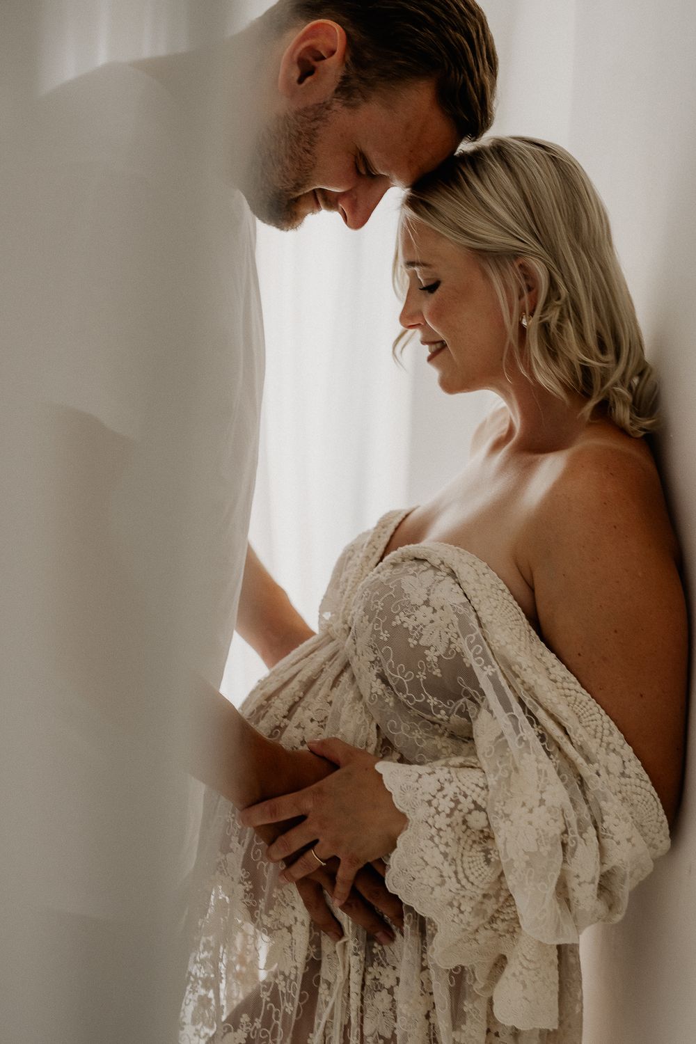 Couple sharing a tender moment, pregnant woman in lace gown cradled by partner near soft white curtains.