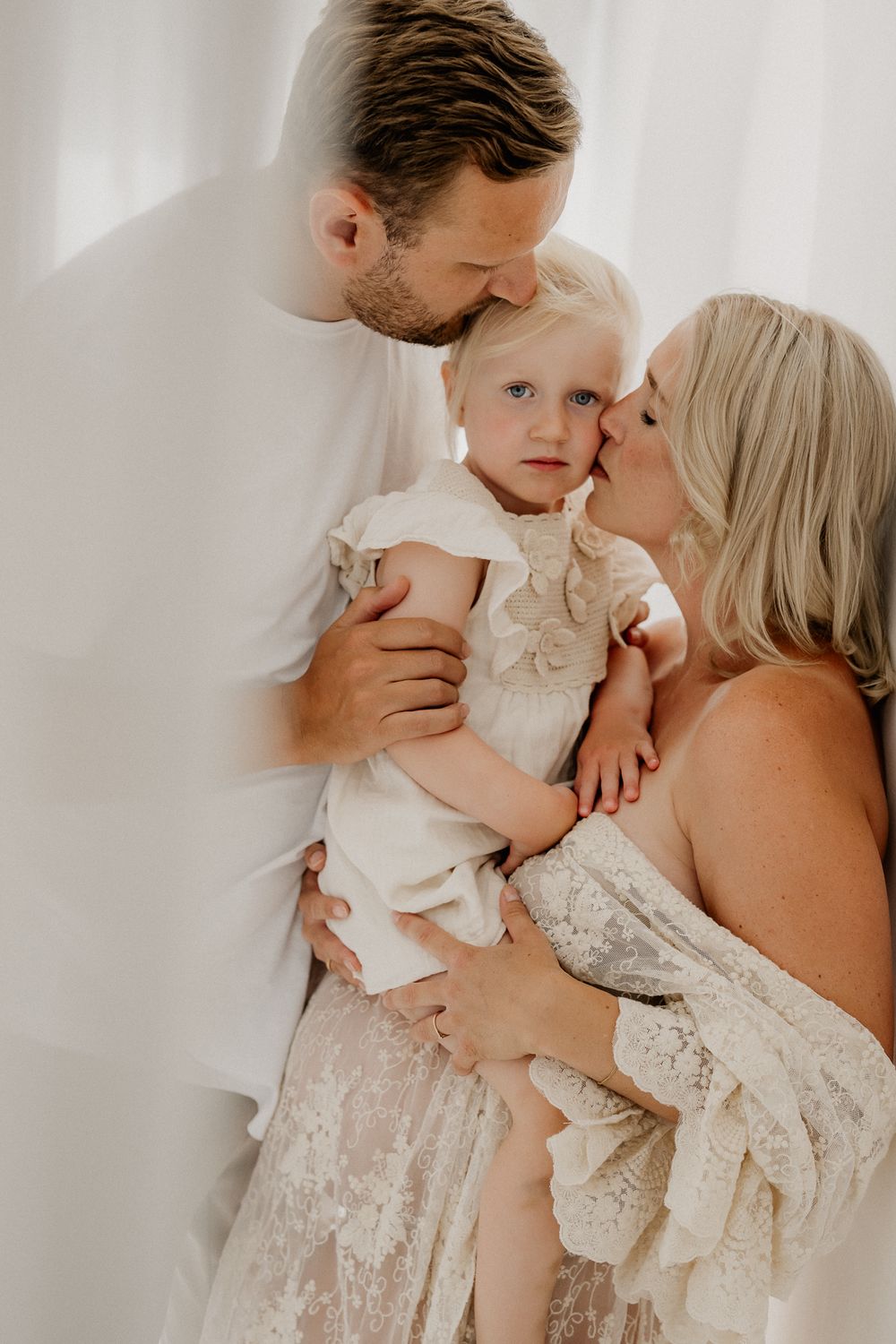 A couple in white outfits lovingly kiss their blonde toddler daughter during a bright, airy family portrait session.