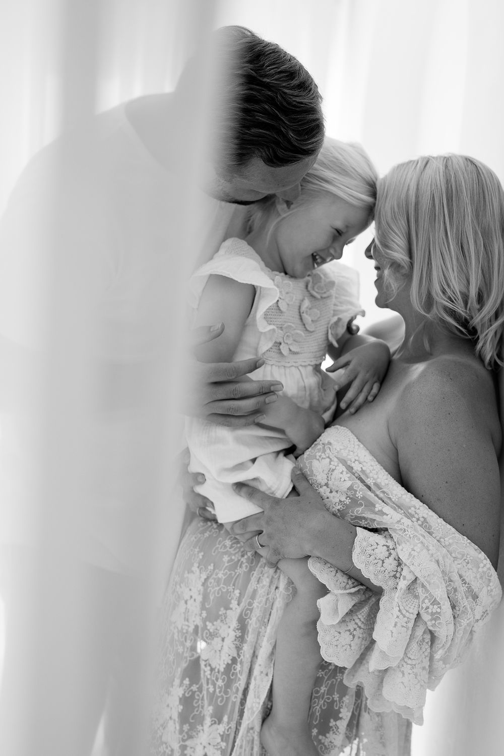 Black and white photo of parents lovingly holding and kissing their smiling baby girl in soft natural light.