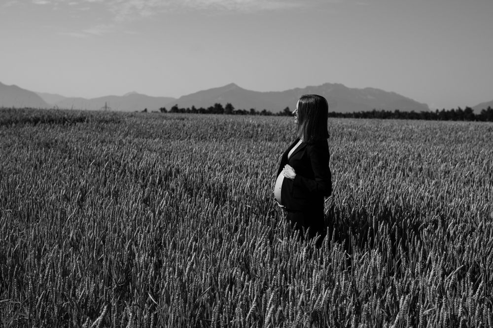 Black and white photo of a pregnant woman standing in a vast wheat field with mountains in the background.