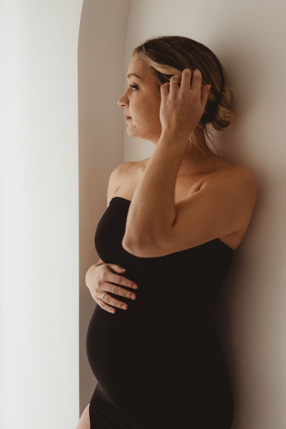 Pregnant woman in black strapless dress leaning against wall, hand in hair, looking to the side.
