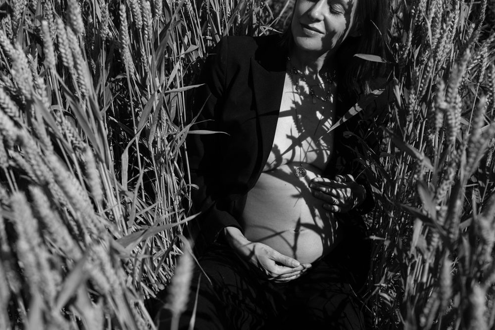 Black and white photo of a pregnant woman sitting in a wheat field, cradling her baby bump in sunlight.