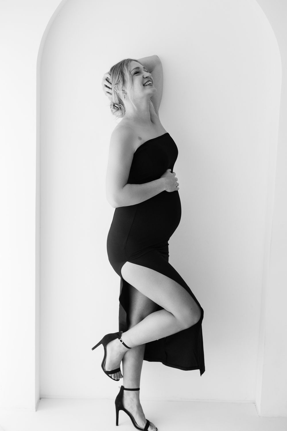 Black and white photo of a smiling pregnant woman in a black strapless dress and heels, cradling her baby bump.