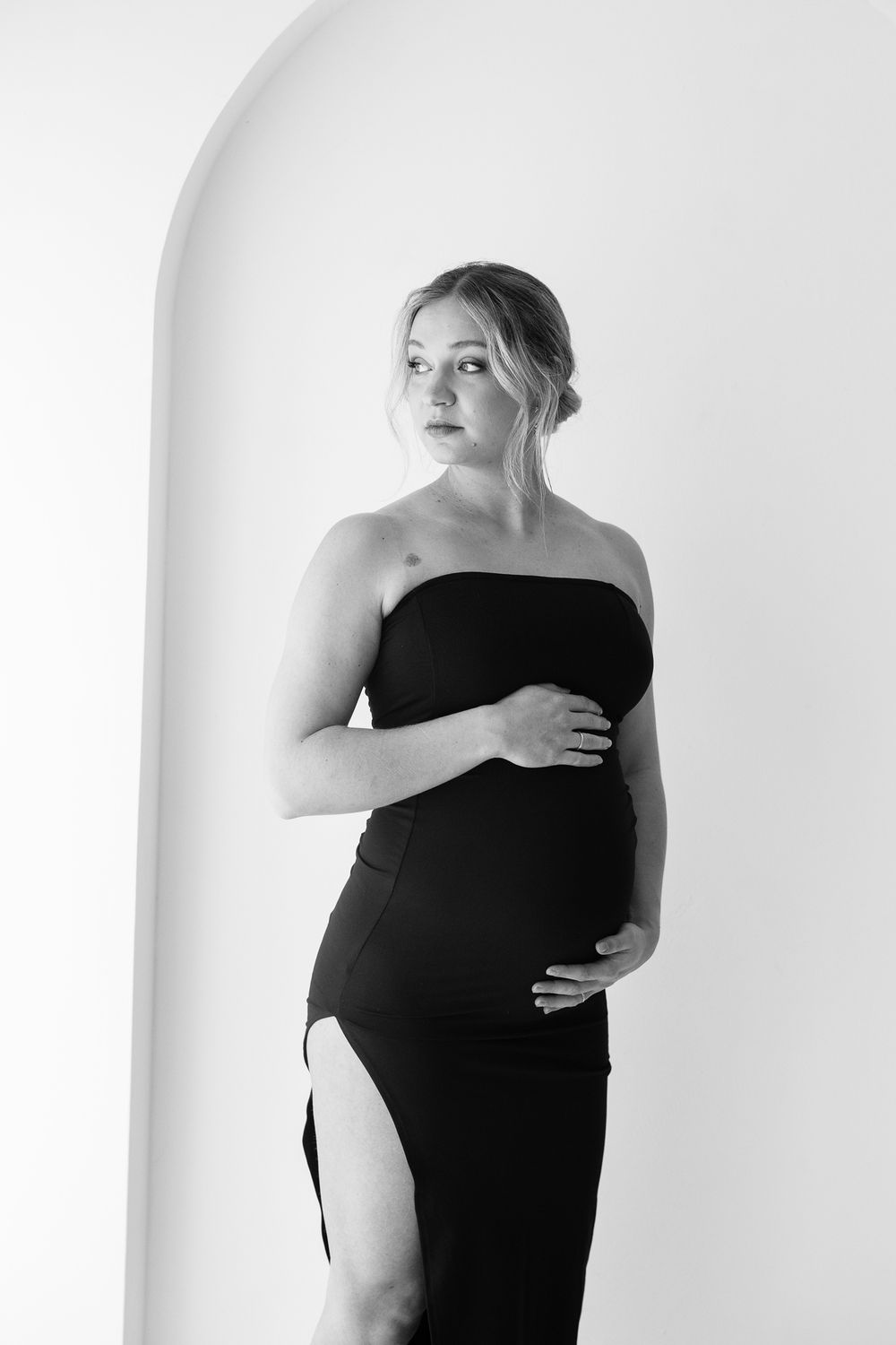 Black and white maternity photo of a blonde woman in a strapless black gown cradling her baby bump near an arched wall.