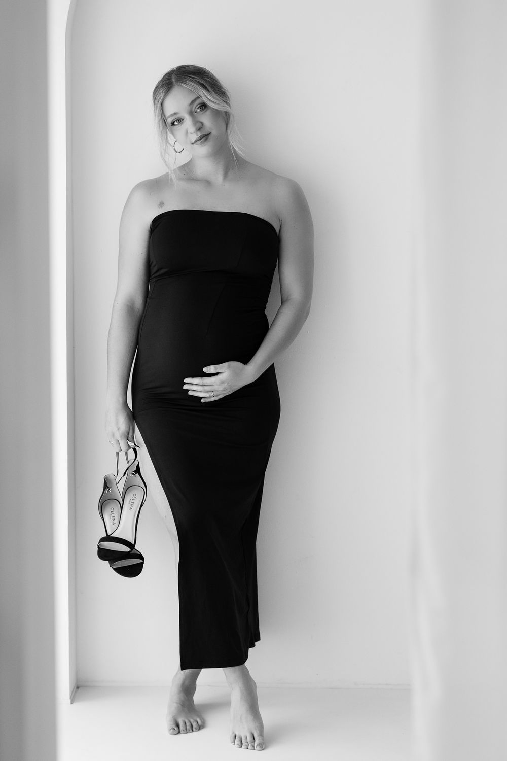 Pregnant woman in black strapless maxi dress holding heels, smiling softly in black and white maternity photo.