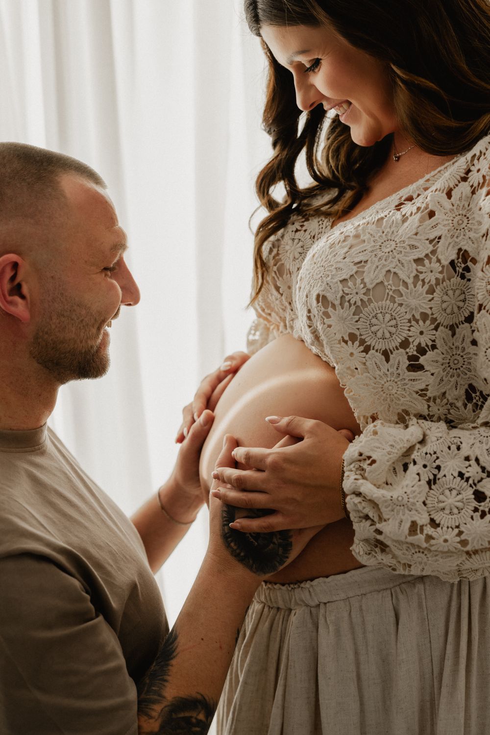 Intimate maternity photo series showing couple in white lace and grey clothing by sheer curtains.