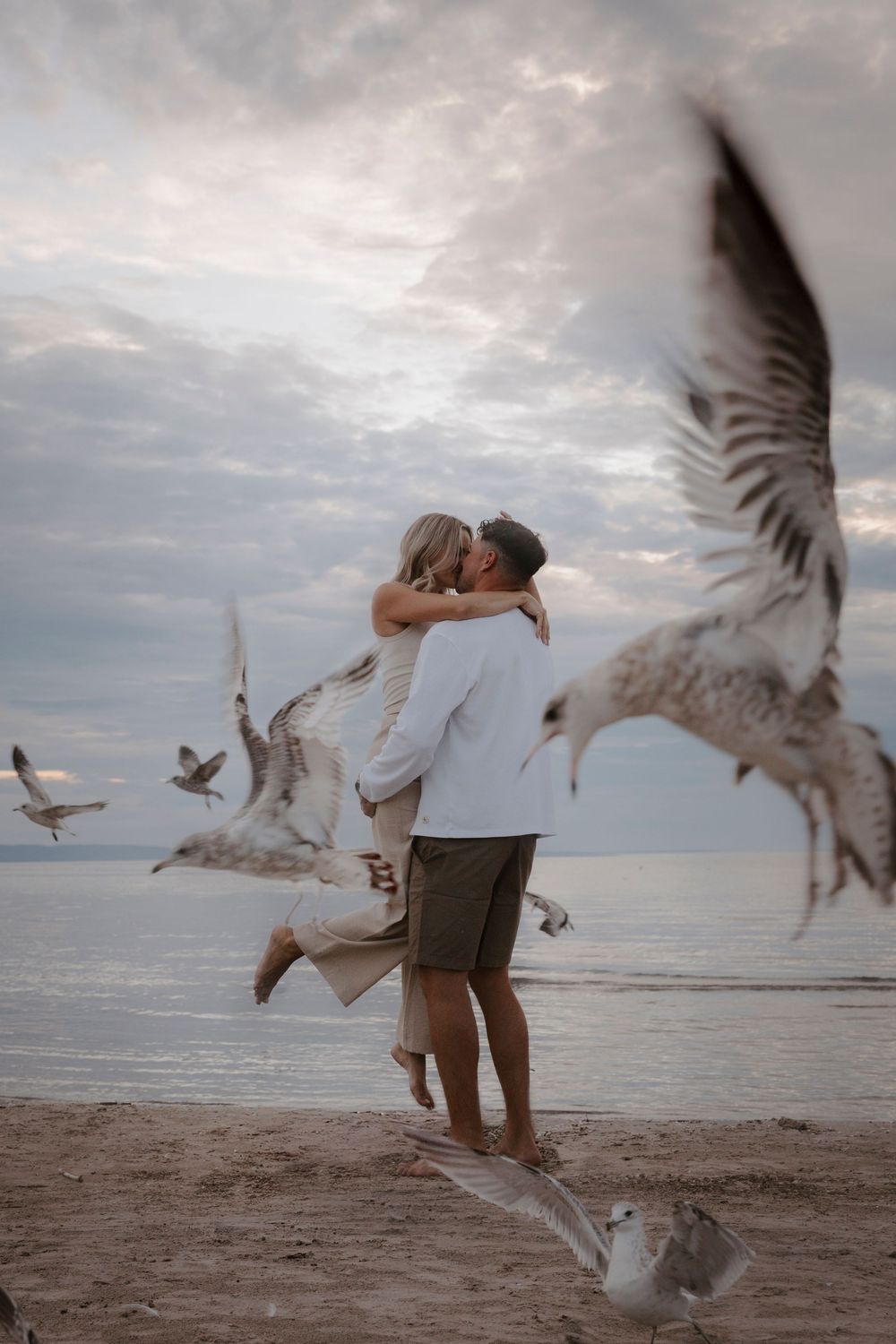 A couple feeds seagulls on a beach at sunset as the birds swoop and dive around them creating a dynamic scene.