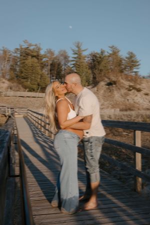 A couple embraces on a wooden pier during sunset with pine trees in the background.