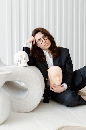 Professional in a black suit sits on the floor with a mannequin head and skull prop against a white backdrop.