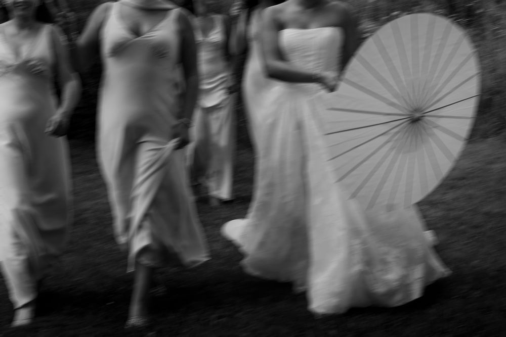 A black and white photograph shows a group of people in white dresses walking with parasols outdoors.