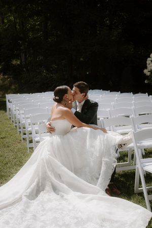 A couple shares a romantic kiss while seated among white folding chairs at an outdoor wedding ceremony.