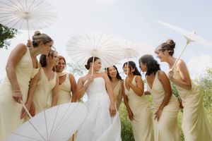 A row of bridesmaids in pale yellow dresses holding white parasols pose together outdoors on a sunny day.