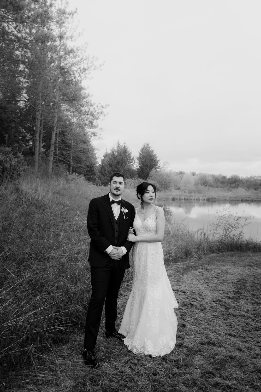 A bride and groom pose together by a lake in a black and white wedding photo.