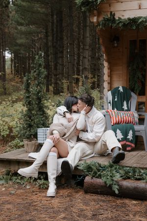 Cozy winter scene with couple in cream outfits sharing intimate moment on rustic cabin porch with holiday decorations.