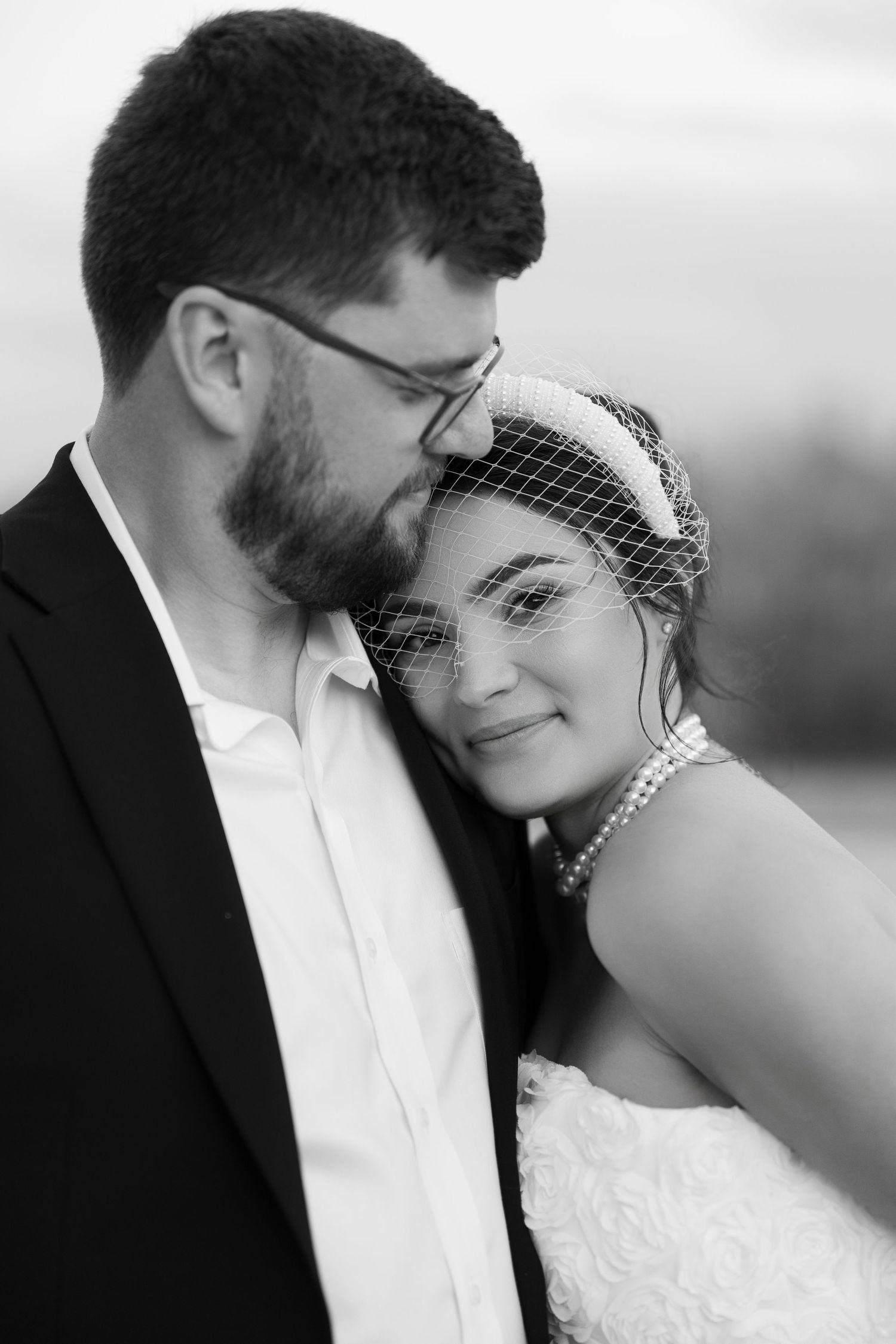 Black and white portrait of bride leaning on groom wearing glasses and dark suit.