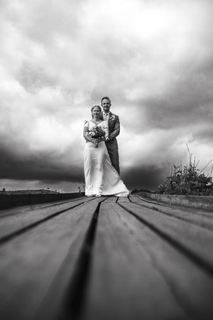 Black and white wedding photo showing a couple standing on wooden boardwalk under dramatic stormy sky.