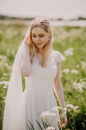 A bride in a white lace wedding dress stands in a meadow of wildflowers on her wedding day.
