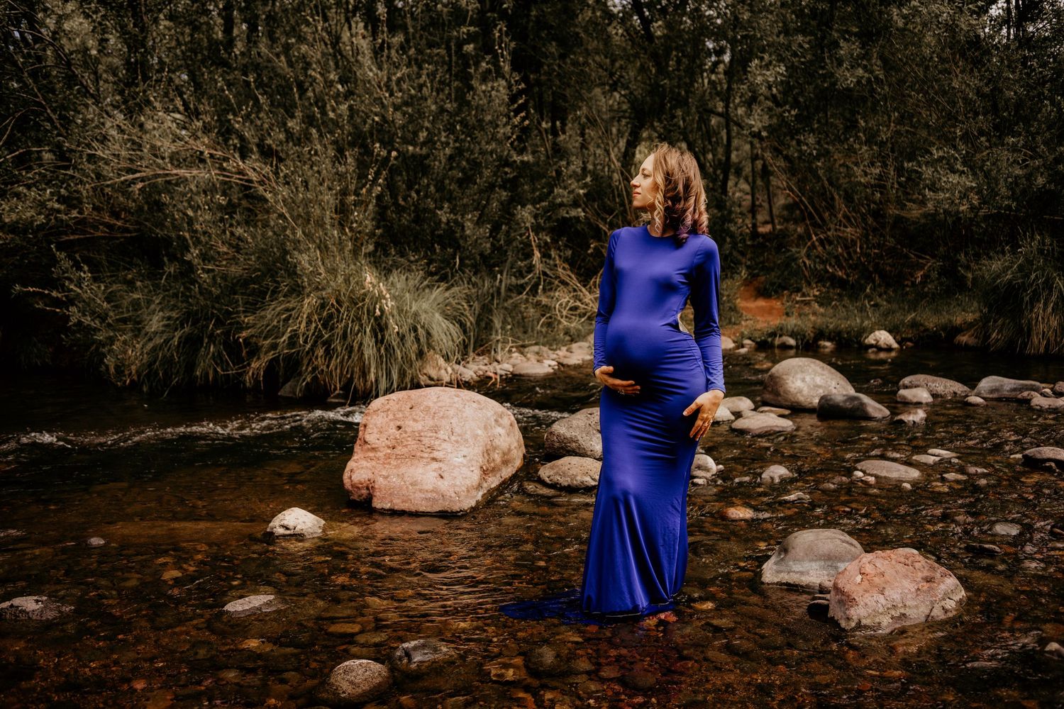 Maternity photo session by a stream featuring a flowing blue dress against rocky natural backdrop.
