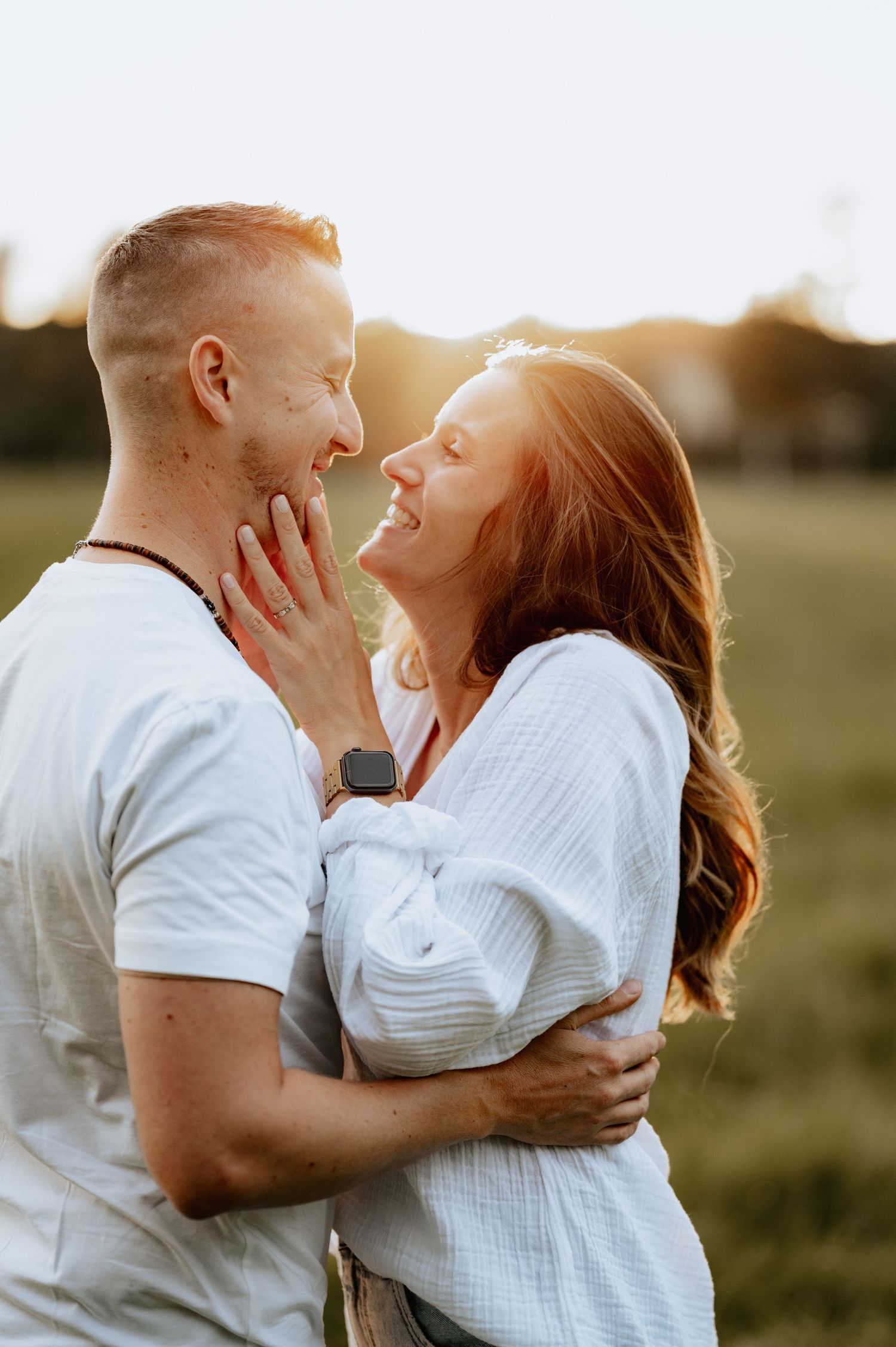 Ein Paar umarmt sich und lacht gemeinsam auf einer sonnigen Wiese während eines romantischen Fotoshootings zur goldenen Stunde.