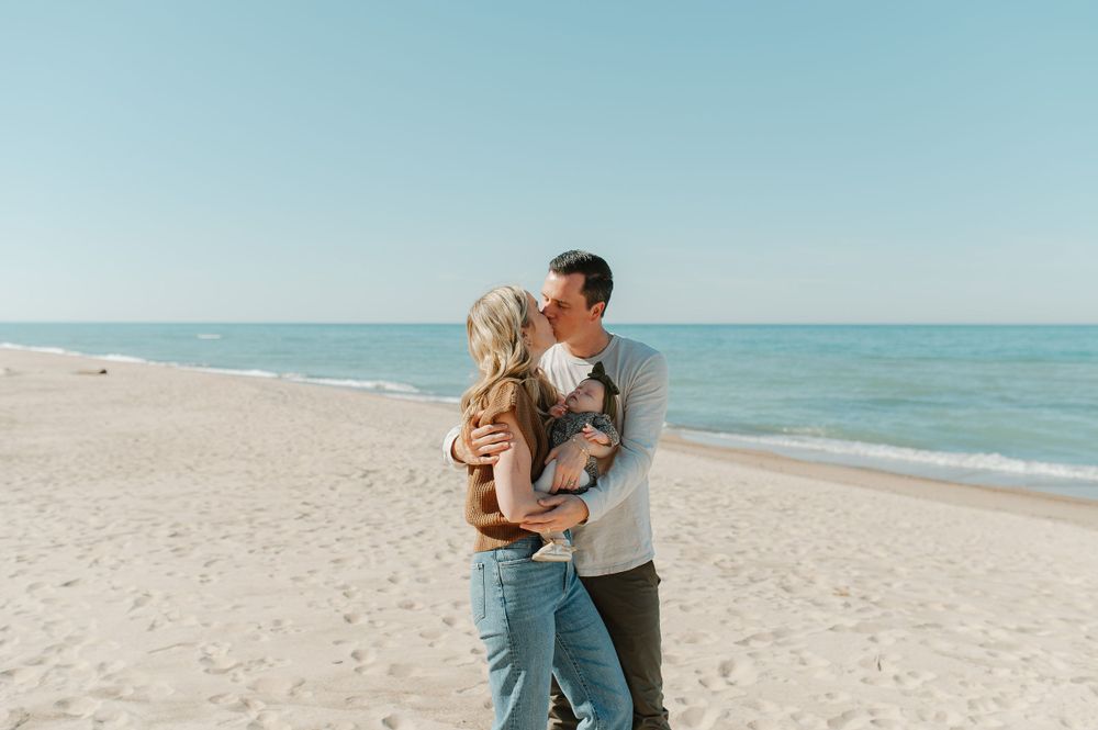 A couple embraces while standing on a sunny beach with turquoise ocean waters in the background.
