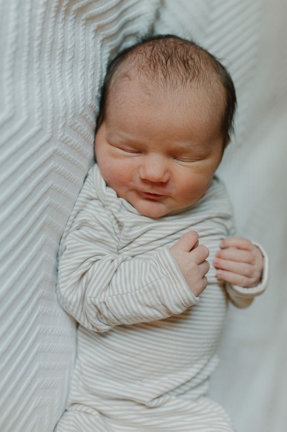 Sleeping newborn baby wearing striped onesie on white textured bedding.
