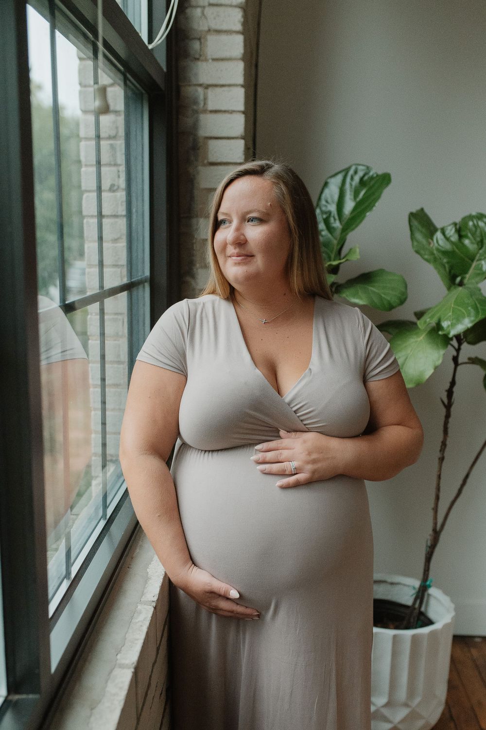 Pregnant woman in grey dress stands by window with indoor plant.
