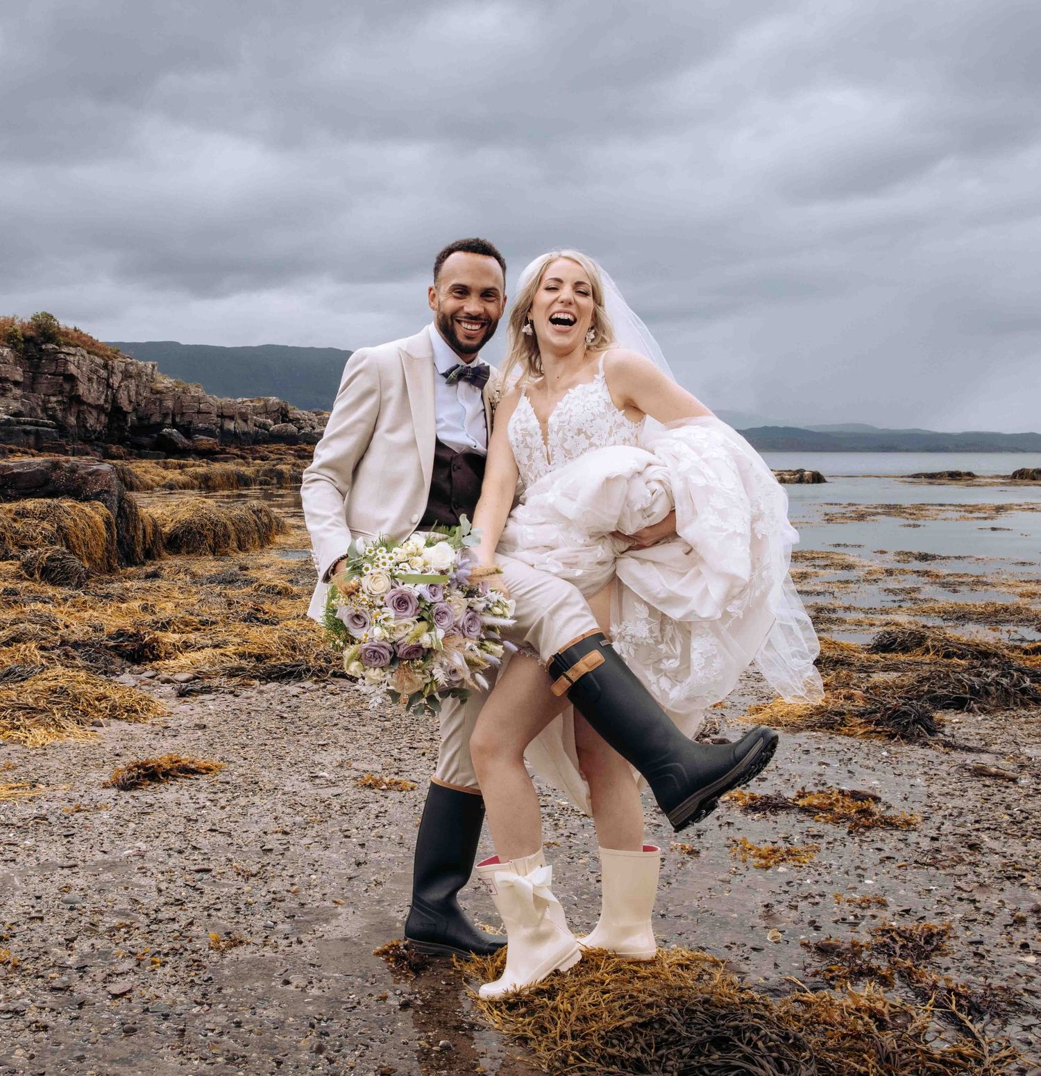 Black and white photo of newlyweds embracing in flowing grass with coastal view.