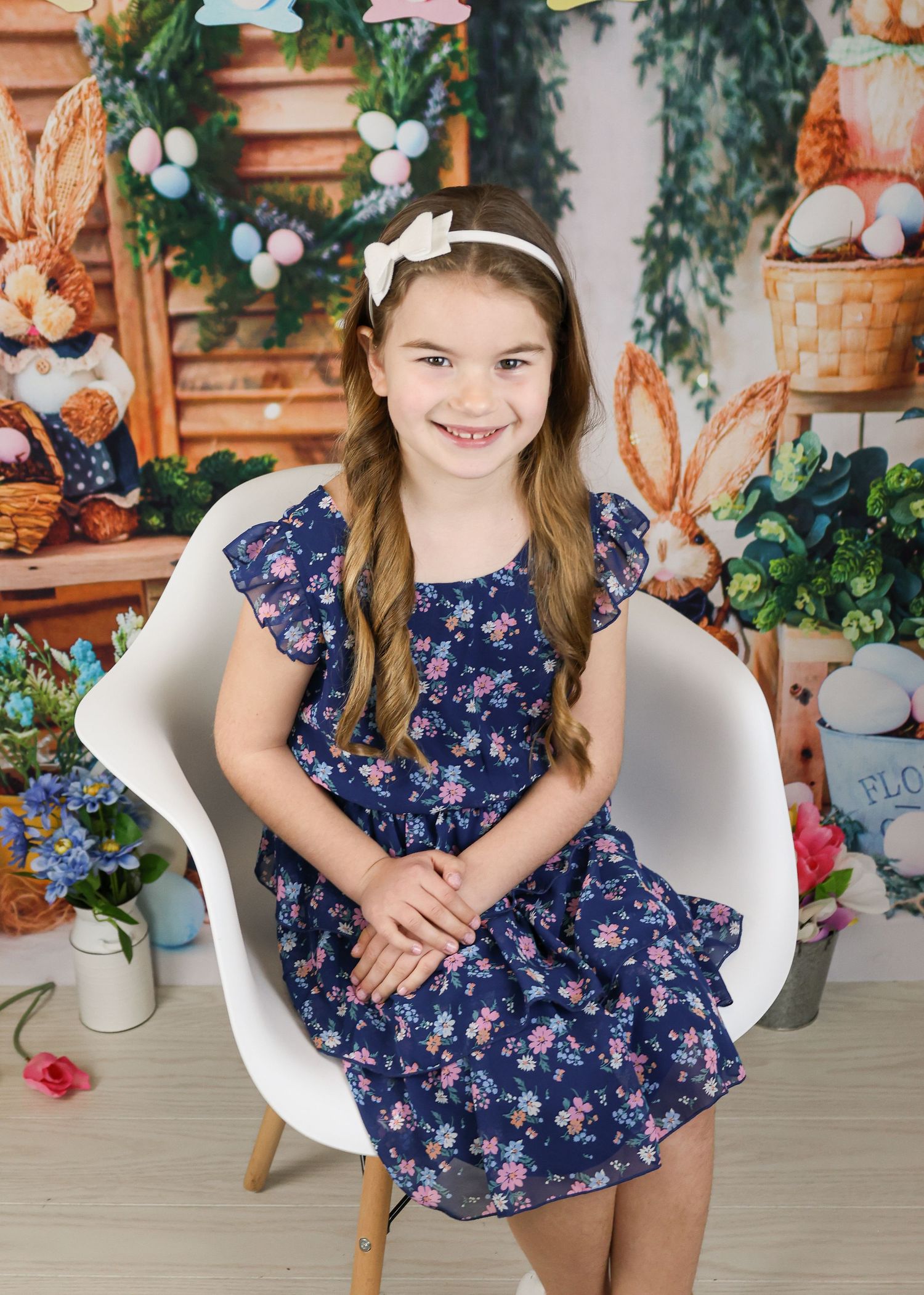Child in navy dress sitting in white chair against spring-themed backdrop with baskets and flowers.