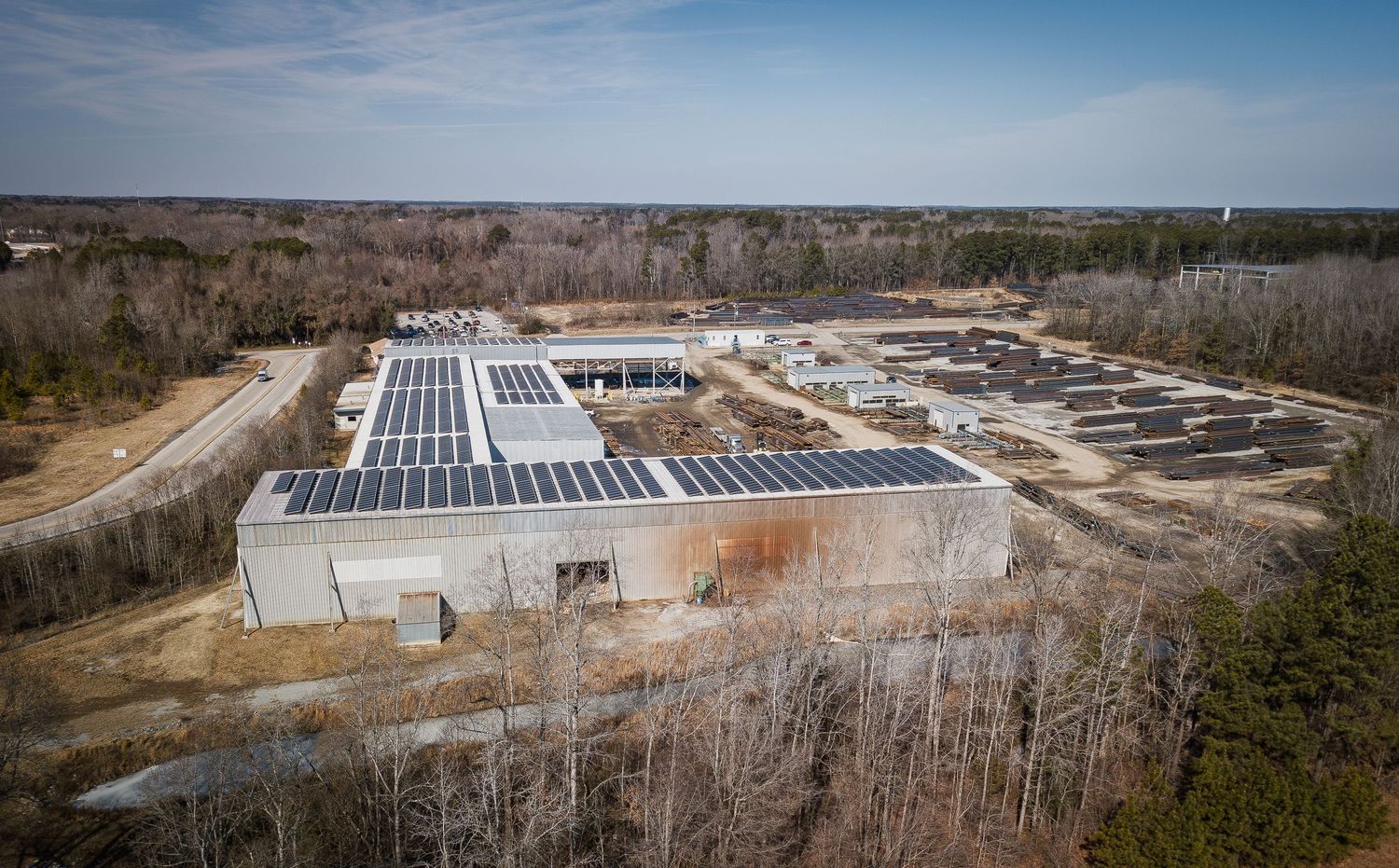 Aerial panoramic view of a large solar panel installation in a rural area surrounded by bare winter trees.