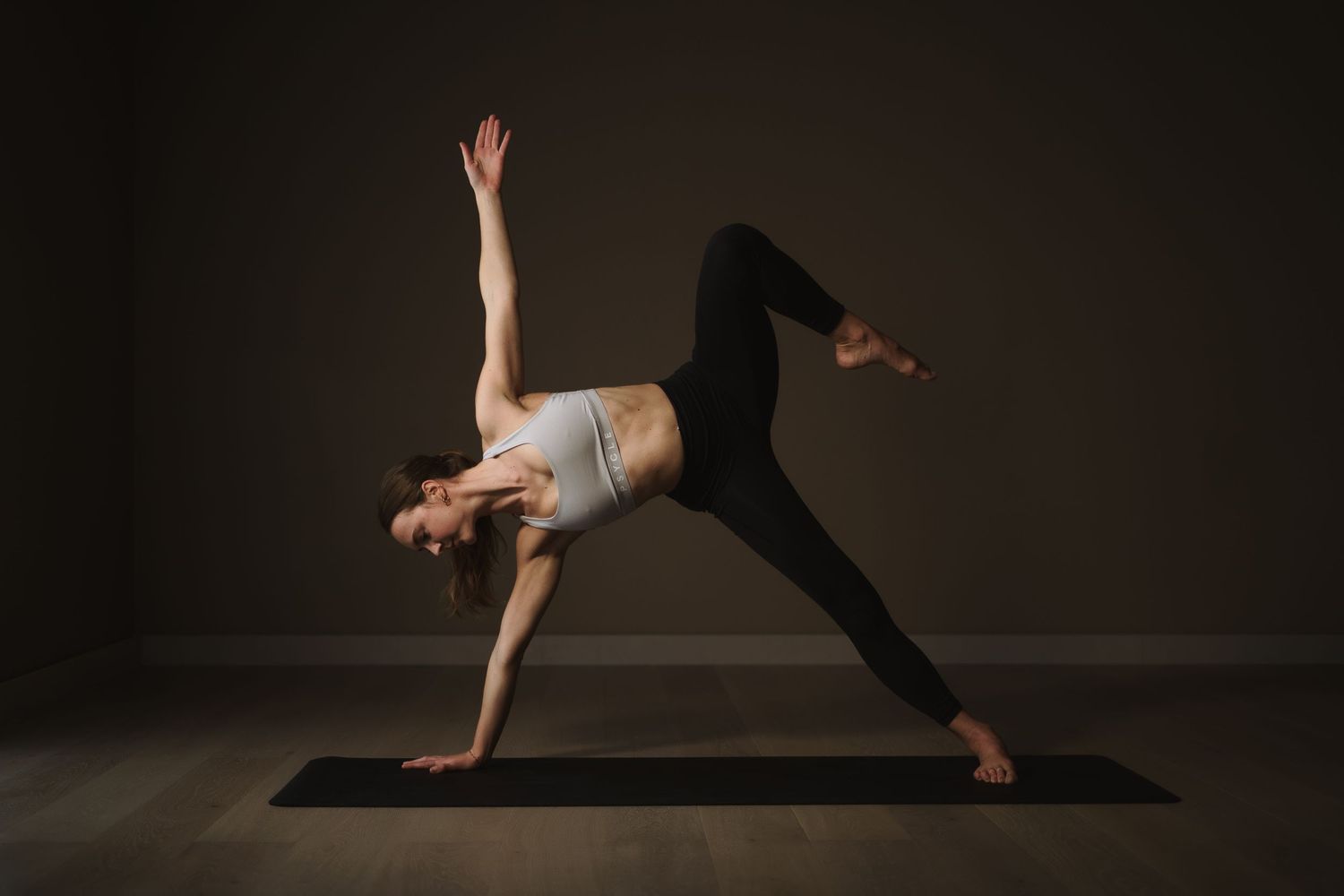 Yoga practitioner demonstrates a dynamic side plank pose on a black mat in a dark studio setting.