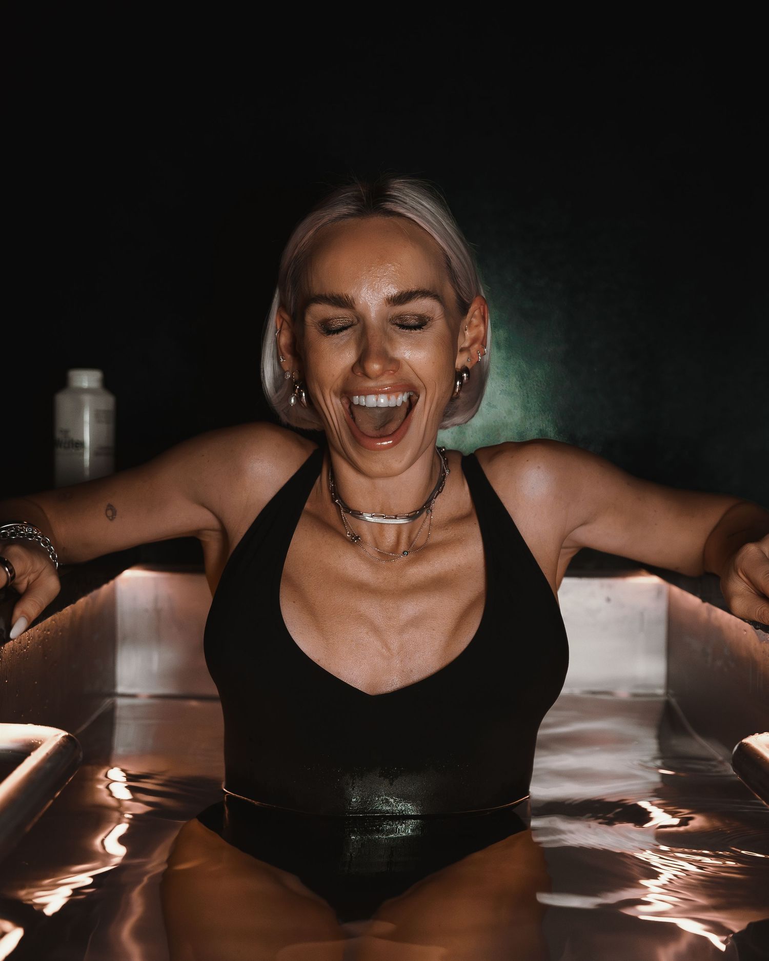 Person wearing black tank top laughing joyfully while posing in dramatic lighting against dark background.
