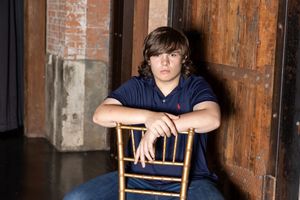 A person sits on a wooden chair against a rustic brick wall in a dimly lit room.