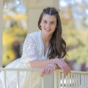 Woman in white lace dress poses outdoors on a porch with autumn foliage in the background.