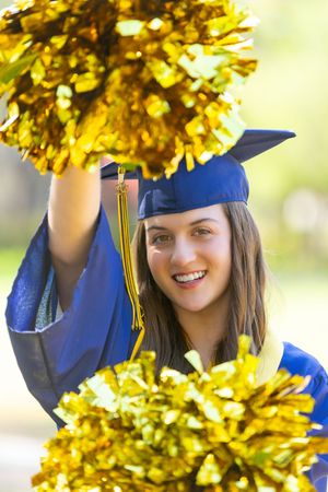 Graduate in blue cap and gown poses with gold pom poms during outdoor graduation photo session.