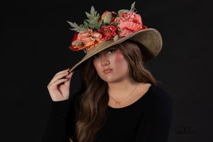 Portrait against dark background wearing a black top and wide-brimmed hat decorated with pink and coral flowers.