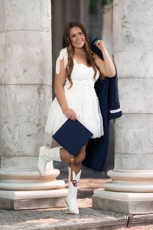 Graduate in white dress and cowboy boots holds diploma and jacket while posing between stone columns.